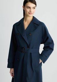 Trench coat blu navy con chiusura a doppio petto, ampio colletto, cintura coordinata, tasche laterali e tessuto dalla consistenza liscia.