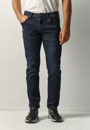 Hombre vestido con jeans slim fit azul oscuro, zapatillas negras y grises, de pie con una mano en el bolsillo y camisa blanca.