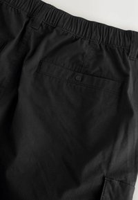 Pantalon à taille élastique noire avec une poche arrière boutonnée et des passants de ceinture sur un tissu léger.