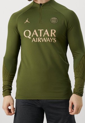 Mężczyzna noszący oliwkowo-zieloną, długą koszulkę treningową PSG z logo Qatar Airways oraz emblematem marki Jordan.