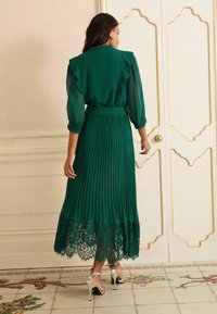 Grünes, plissiertes Kleid mit Spitze am Saum, Rüsche an den Schultern, transparenten langen Ärmeln und taillierter Taille. Das Model trägt silberne High Heels.