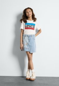 Bijela majica s crnim i crvenim Levi's logotipom, svijetloplava traper suknja s cvjetnim vezom i bež visoke tenisice s bijelim čarapama.