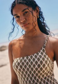 Jednoroční plavky v krémové barvě se zeleným a zlatým listovým vzorem, s detaily pleteného ramínka. Hladká textura, přiléhavý střih.