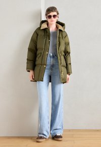 Jachetă puffer de culoare verde măslinie cu guler crem, asortată cu blugi largi de culoare albastru deschis și pantofi cu platformă de culoare maro. Material texturat, buzunare frontale.