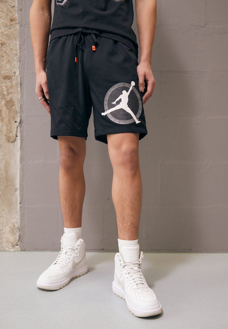 Personne portant un short noir Air Jordan avec le logo Jumpman et des baskets montantes blanches Nike, faisant face à un mur gris.