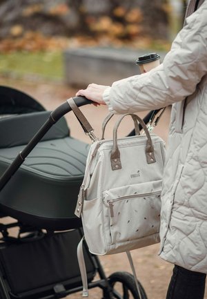 Person wearing a quilted jacket holding a stroller handle with a beige bag attached and a takeaway coffee cup in the background.