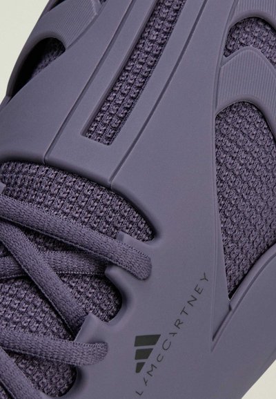 Close-up of purple sneaker with knit fabric, rubber overlays, laces, and logo text "Stella McCartney" on the side.