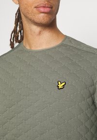 T-shirt acolto verde oliva realizzato in misto cotone, con scollatura rotonda e un piccolo logo di un uccello ricamato giallo sul petto.