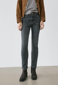 Personne portant un jean slim gris foncé, une veste en daim marron, une chemise grise et des bottines en daim marron foncé, debout sur un sol clair.