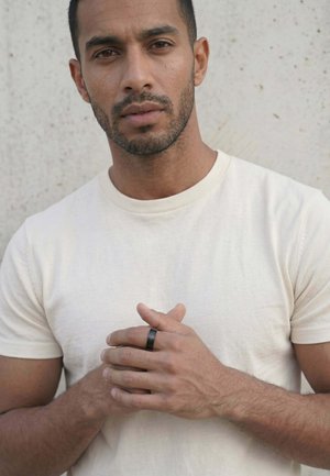 Uomo con capelli corti e scuri e barba, che indossa una maglietta bianca, con le mani intrecciate che mostrano un anello nero al dito su uno sfondo chiaro e strutturato.