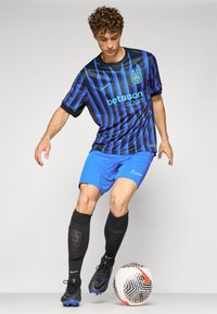 Nike jalkapallovarustus, joka sisältää musta-siniraidallisen paidan, siniset shortsit ja mustat puristussukat, yhdistettynä kuviolliseen jalkapalloon.