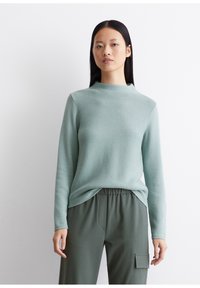 Mintgrüner Pullover mit hohem Kragen und langen Ärmeln, ausgestattet mit einem strukturierten Strickmuster. Kombiniert mit olivgrünen, elastischen Hosen mit Taillenbund.