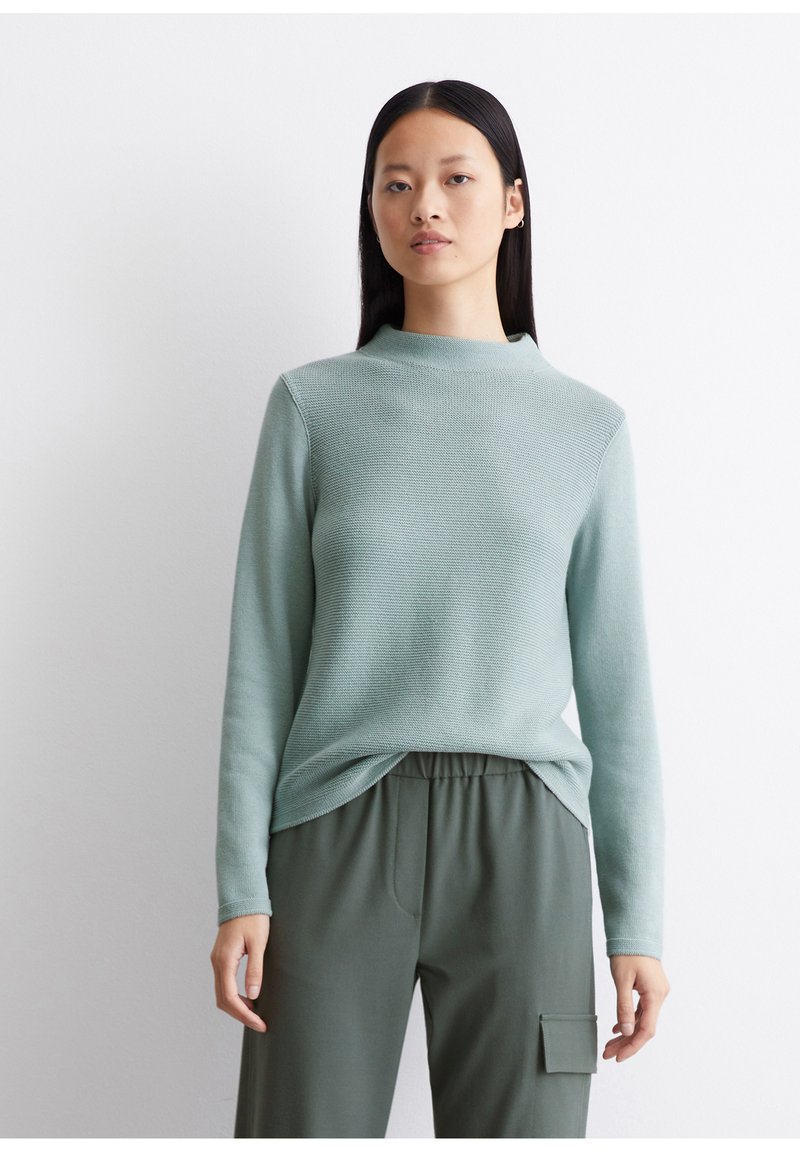 Mintgrüner Pullover mit hohem Kragen und langen Ärmeln, ausgestattet mit einem strukturierten Strickmuster. Kombiniert mit olivgrünen, elastischen Hosen mit Taillenbund.