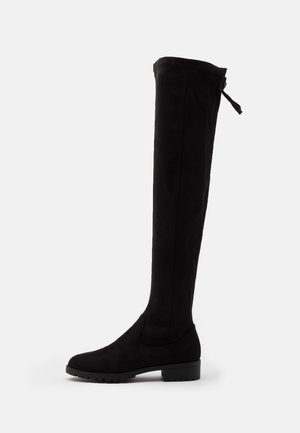 Botte plate noire montante au-dessus du genou, avec bout arrondi, talon bas, semelle texturée et détail de sangle ajustable en haut à l'arrière.
