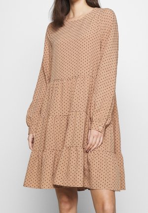 Beiges langärmeliges Kleid mit schwarzen Punkten, Stufenrock und geknöpften Manschetten, getragen von einer stehenden Frau vor weißem Hintergrund.