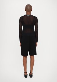 Black long-sleeve top with sheer fabric, layered over black shorts. Features adjustable straps and a smooth texture. Black footwear with studs.