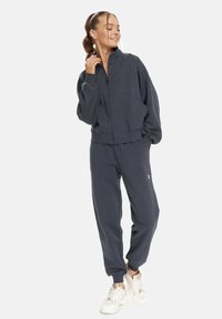 Marineblauer Reißverschluss-Sweatshirt mit hohem Kragen, lockerer Passform und geripptem Saum, kombiniert mit passenden Jogginghosen. Weißes Logo am linken Bein.
