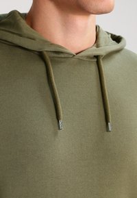 Sudadera verde oliva hecha de material suave, con una gran capucha y cordones ajustables con extremos metálicos. Textura suave en todo su alrededor.
