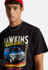 Černé tričko s grafickou potiskem modrého dodávky a nápisem "HAWKINS Hitmaker!" v výrazných barvách. Krátké rukávy, žebrovaný límec.