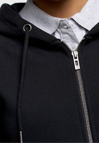 Felpa nera con zip e camicia grigia con colletto sotto. Presenta una cerniera argentata, lacci e una consistenza morbida. Inquadratura ravvicinata del collo.
