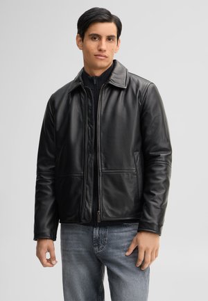 Junger Mann trägt eine schwarze Lederjacke über einem schwarzen Zip-Pullover und blauen Jeans, steht vor einem schlichten grauen Hintergrund.