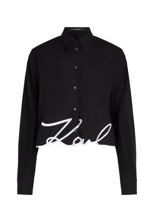 Camisa negra corta con mangas largas, botón frontal y un gran logo bordado en blanco "Karl" en el dobladillo. Tela suave y lisa.