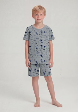 Garçon blond debout pieds nus portant un short gris assorti et un T-shirt avec des imprimés bleu marine sur le thème de l’espace, astronaute et fusée.