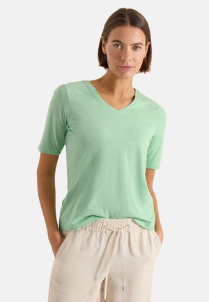 Kvinde med kort brunt hår iført en mintgrøn V-hals kortærmet skjorte og beige bukser med snøre i taljen, med hænderne i lommerne.