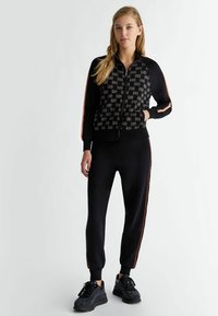 Giacca nera con zip e davanti a fantasia, con strisce laterali rosse a contrasto, abbinata a pantaloni neri e sneakers nere strutturate.