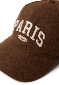 Bruine wollen pet met "PARIS" in geborduurde witte letters, een ronde klep en textiel met structuur. Verdere details omvatten een "91"-label.