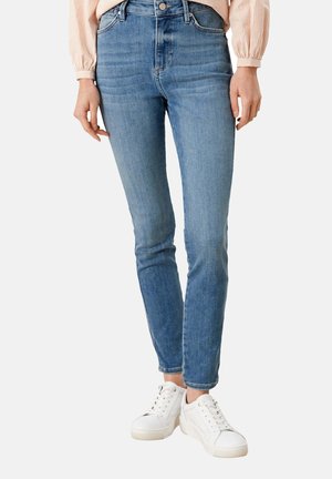 Mujer que lleva jeans ajustados azul claro, blusa beige con mangas remangadas y zapatillas blancas, de pie contra un fondo liso.