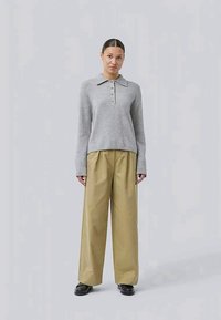 Donna in piedi che indossa un maglione grigio a maniche lunghe con colletto e bottoni, pantaloni beige larghi e scarpe nere su uno sfondo chiaro e uniforme.
