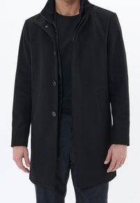 Musta villakangastakki korkealla kauluksella, etupainikkeilla ja sivutaskuilla; sileä pinta; yhdistetty mustan paidan ja tummakuvioisten housujen kanssa.