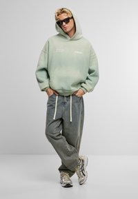 Sudadera verde menta con bolsillo frontal y texto blanco, combinada con vaqueros de mezclilla ligera y sueltos, y zapatillas elegantes, presentada en un entorno minimalista.