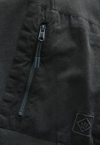 Juoda audinio medžiaga su tekstūruotu paviršiumi, turinti kišenę su užtrauktuku ir logotipo pleistrą apatiniame kampe. Medžiaga turi matinį atspalvį.