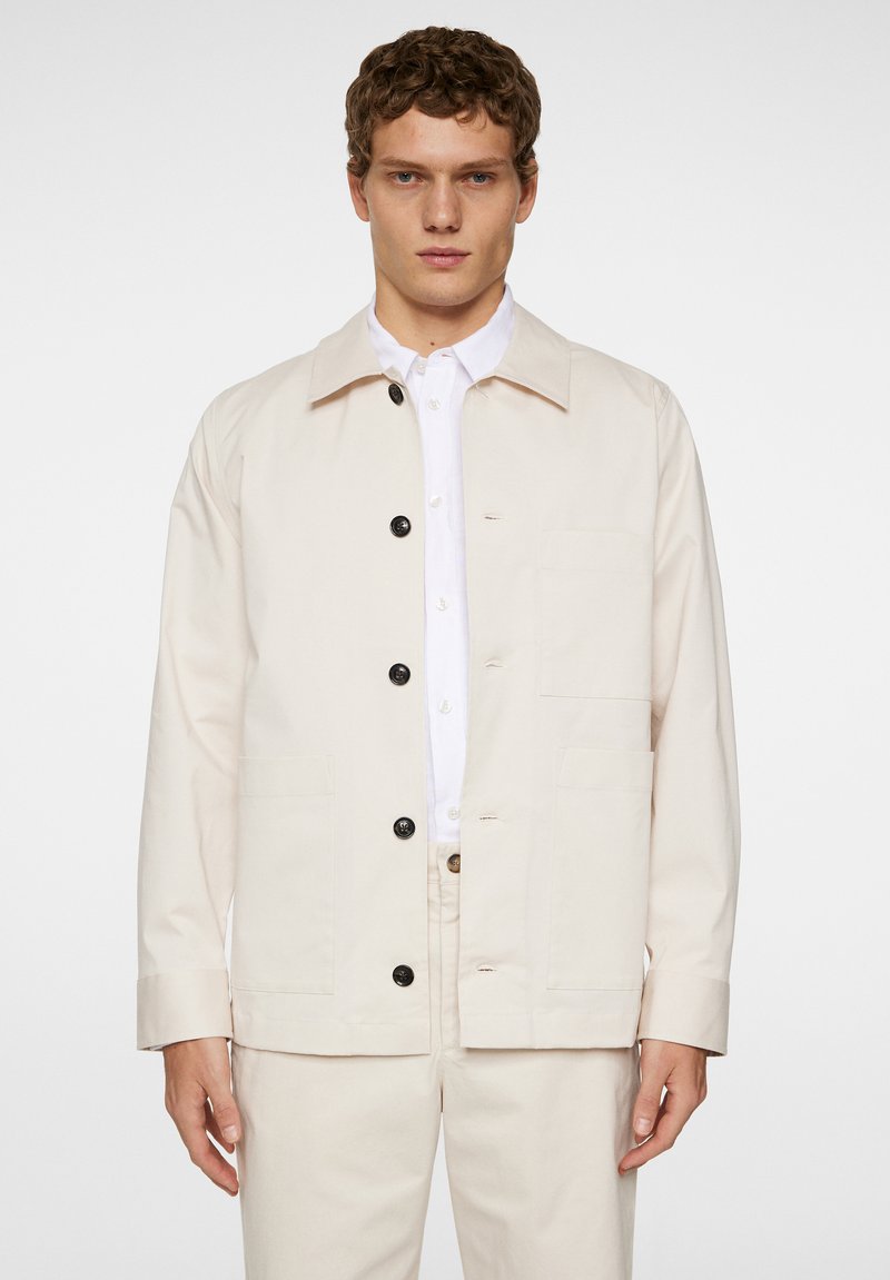 Lichtbeige jasje met een rechte snit, knoopsluiting en twee voorkorre pockets. Gemaakt van gladde stof met een strakke kraag.