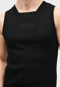 Μαύρο αμάνικο ριμπ μπλουζάκι με τετράγωνο ντεκολτέ και κεντημένο το "Versace Jeans Couture" στο στήθος, φορεμένο από ένα άτομο.