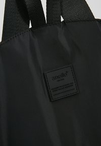 Tissu de sac à dos noir avec deux bretelles et une étiquette de marque noire cousue indiquant « anello » ainsi qu'un texte supplémentaire en petit.