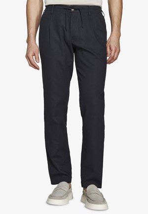 Mężczyzna w czarnych casualowych spodniach z sznurkiem w pasie i beżowych butach na wsuwkę, stojący na białym tle.