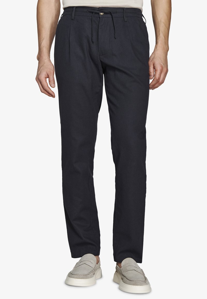 Hombre con pantalones casuales negros con cintura ajustable y zapatos beige sin cordones, de pie contra un fondo blanco.