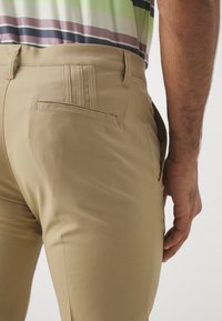 Pantalones caqui de corte ajustado, con un bolsillo trasero plano, pasadores para cinturón y una textura de tela suave. Parte superior a rayas de varios colores visible arriba.