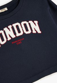 Sudadera azul marino con la palabra "LONDRES" en letras blancas texturizadas con borde rojo, y "Athletic&Leisure 1985" bordado abajo.