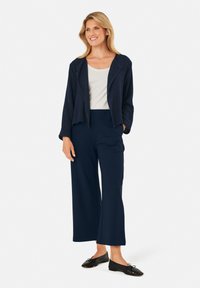 Marineblauwe cropped jacket met een zachte textuur, gedragen over een lichte top en gecombineerd met brede marineblauwe broeken; afgemaakt met zwarte ballerina's.