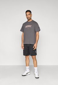 Szara koszulka z krótkim rękawem z nadrukiem graficznym, zestawiona z czarnymi szortami i białymi butami sportowymi z czarnym akcentem. Prosty, casualowy styl.