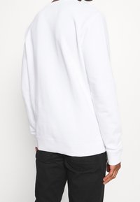 Vit sweatshirt i mjukt material, med långa ärmar och rundad halsringning. Den nedre fållen är något ribbad, kombinerad med mörka byxor.