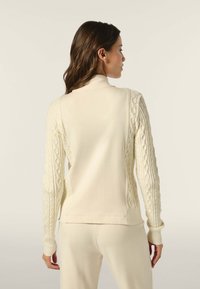 Maglione color crema con maniche a treccia, collo alto e polsini a costine. Presenta un corpo liscio e spaccature laterali all'orlo.