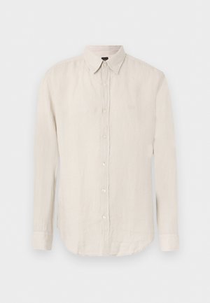 Cămașă bej cu mâneci lungi și nasturi, cu guler ascuțit și logo subtil pe pieptul stâng, prezentată pe un fundal simplu.