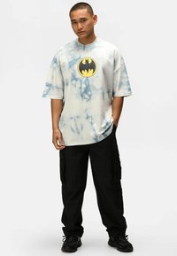 Camiseta de algodón de color azul claro con diseño de tie-dye, con cuello redondo y un gran logo de Batman en negro y amarillo en la parte frontal, combinada con pantalones cargo negros.