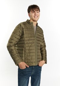 Giacca puffer verde oliva con design a righe verticali, chiusura con zip frontale e collo alto, indossata sopra un maglione grigio, abbinata a jeans blu.