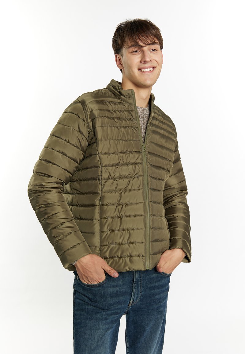 Giacca puffer verde oliva con design a righe verticali, chiusura con zip frontale e collo alto, indossata sopra un maglione grigio, abbinata a jeans blu.